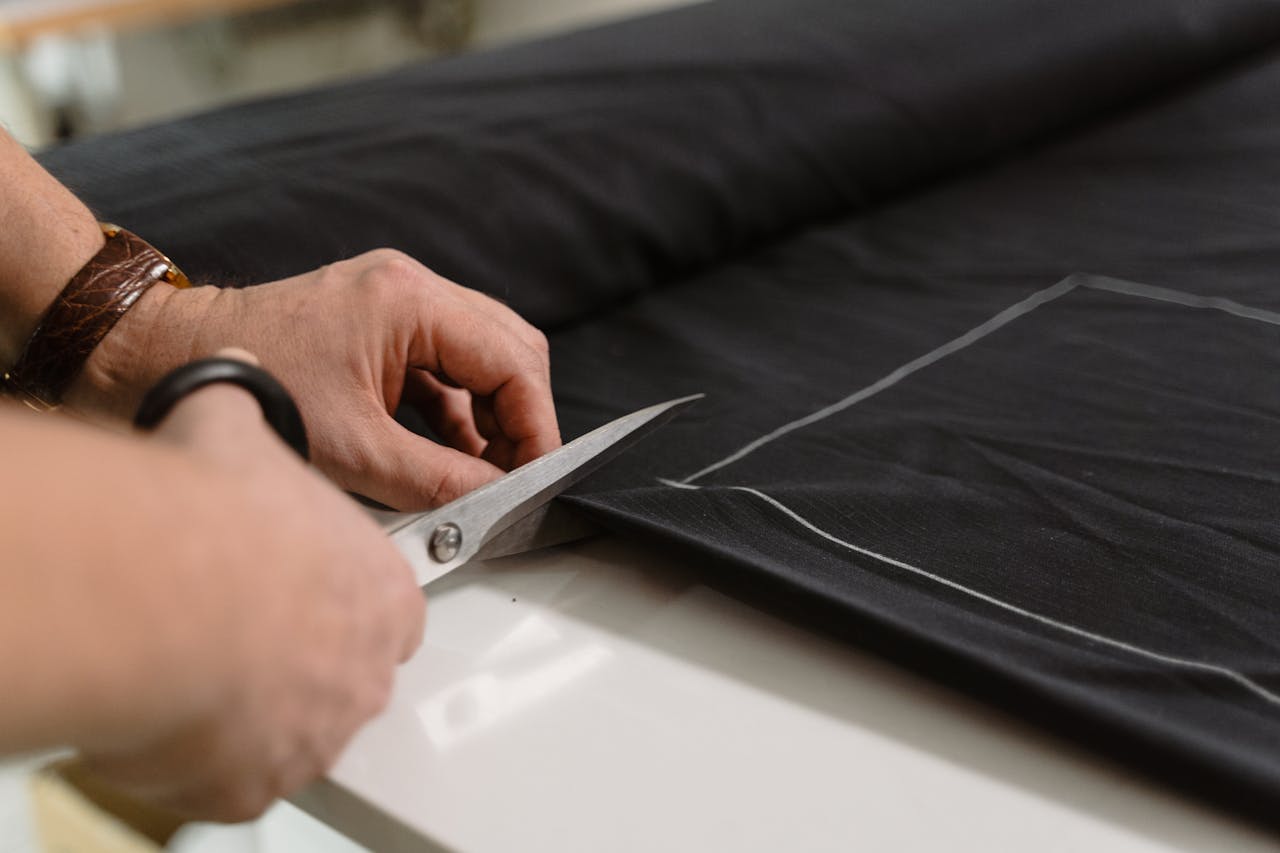 Services Close-up of a tailor's hand cutting black fabric with scissors, highlighting craftsmanship.